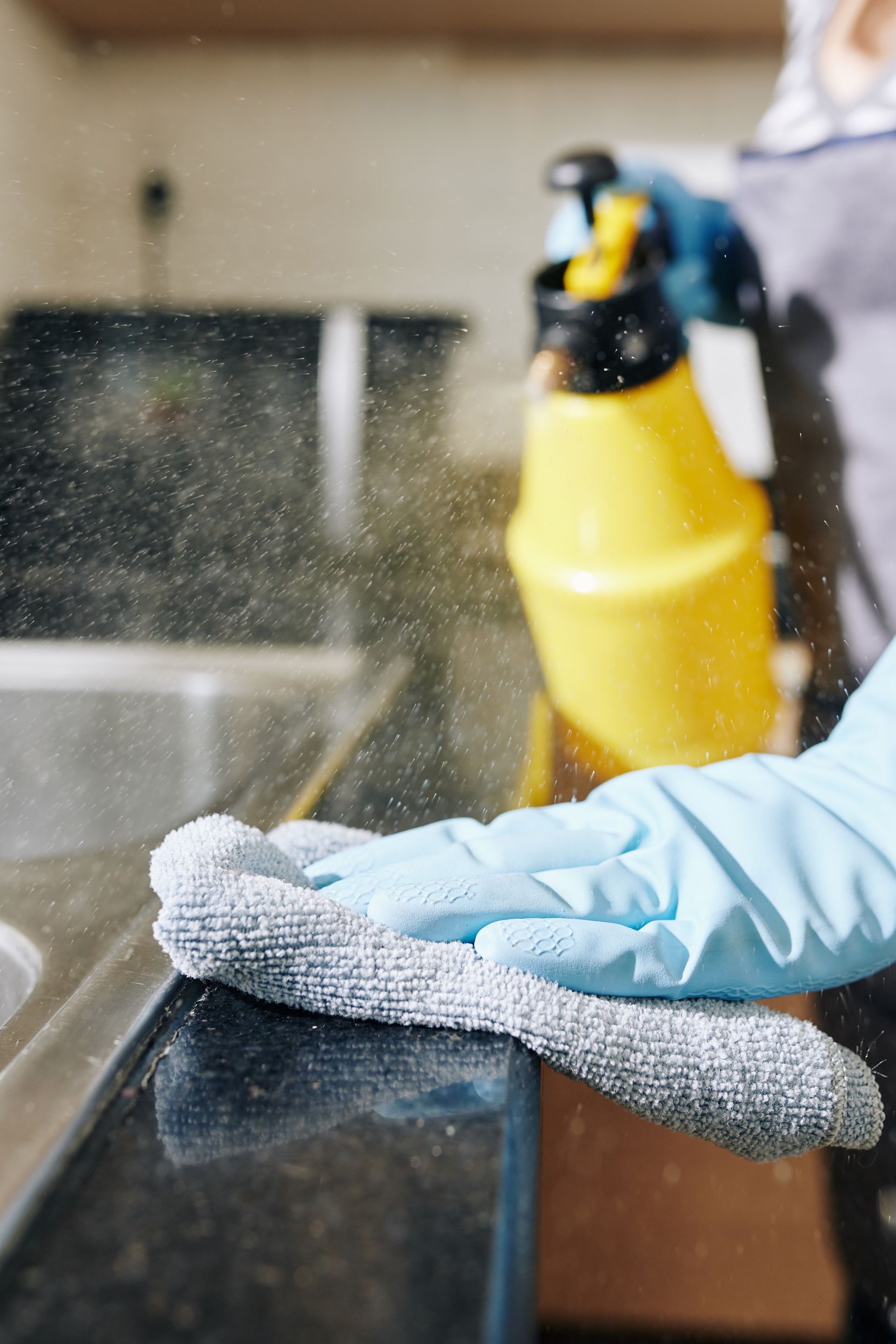 Gloved hand wiping a black countertop with a gray cloth, spray bottle in the background