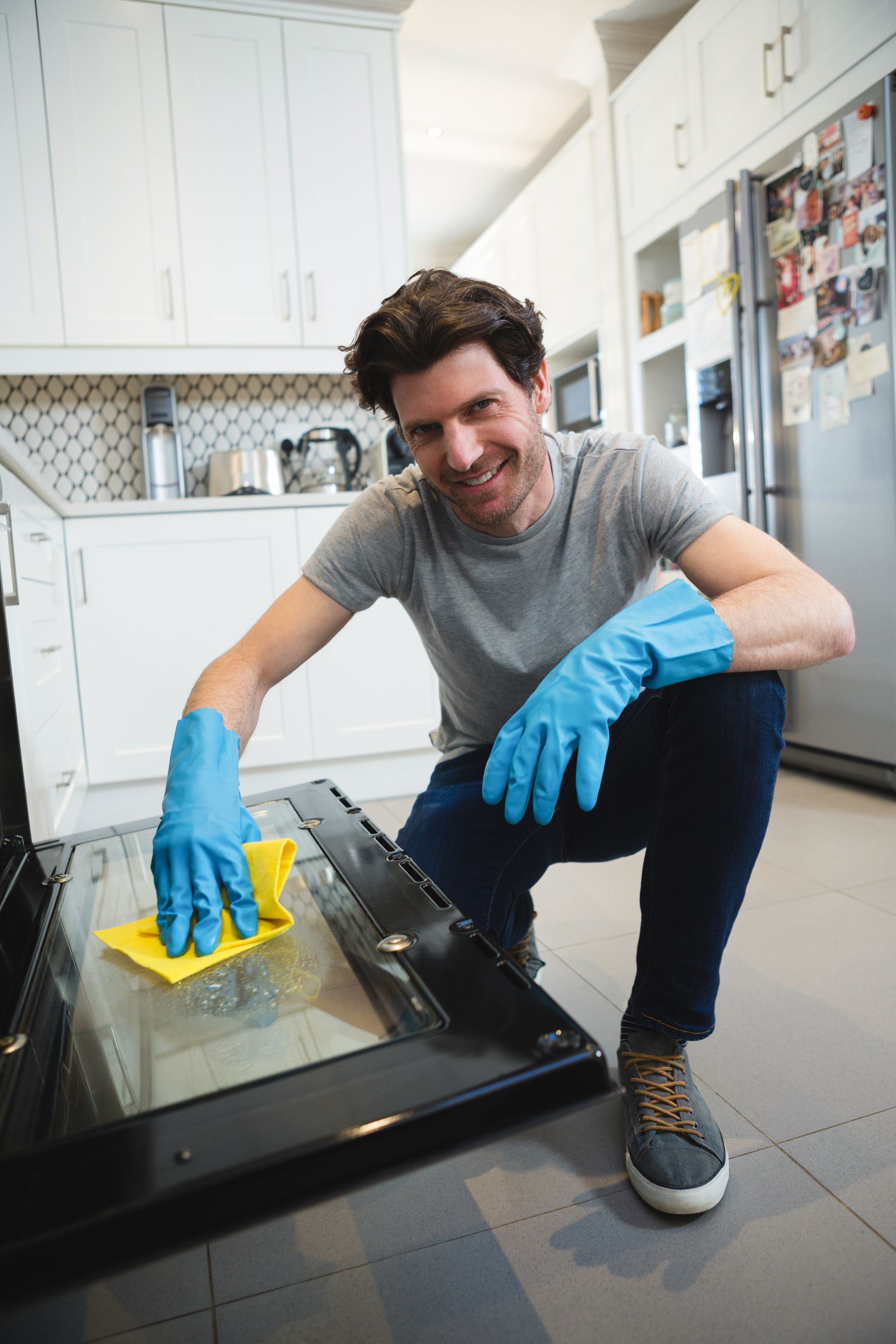 Man cleaning a glass oven door in a bright kitchen, wearing blue gloves and smiling.