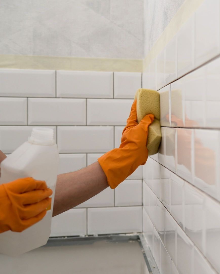 Gloved hands cleaning white subway tile wall with a sponge in a tiled room