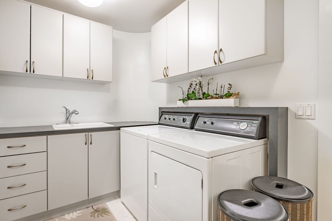 Bright white laundry room with stacked washer and dryer, cabinets, countertop, and a small plant shelf.