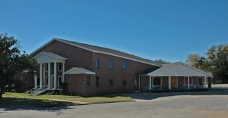 Brick building with white columns and a covered porch on a sunny day.