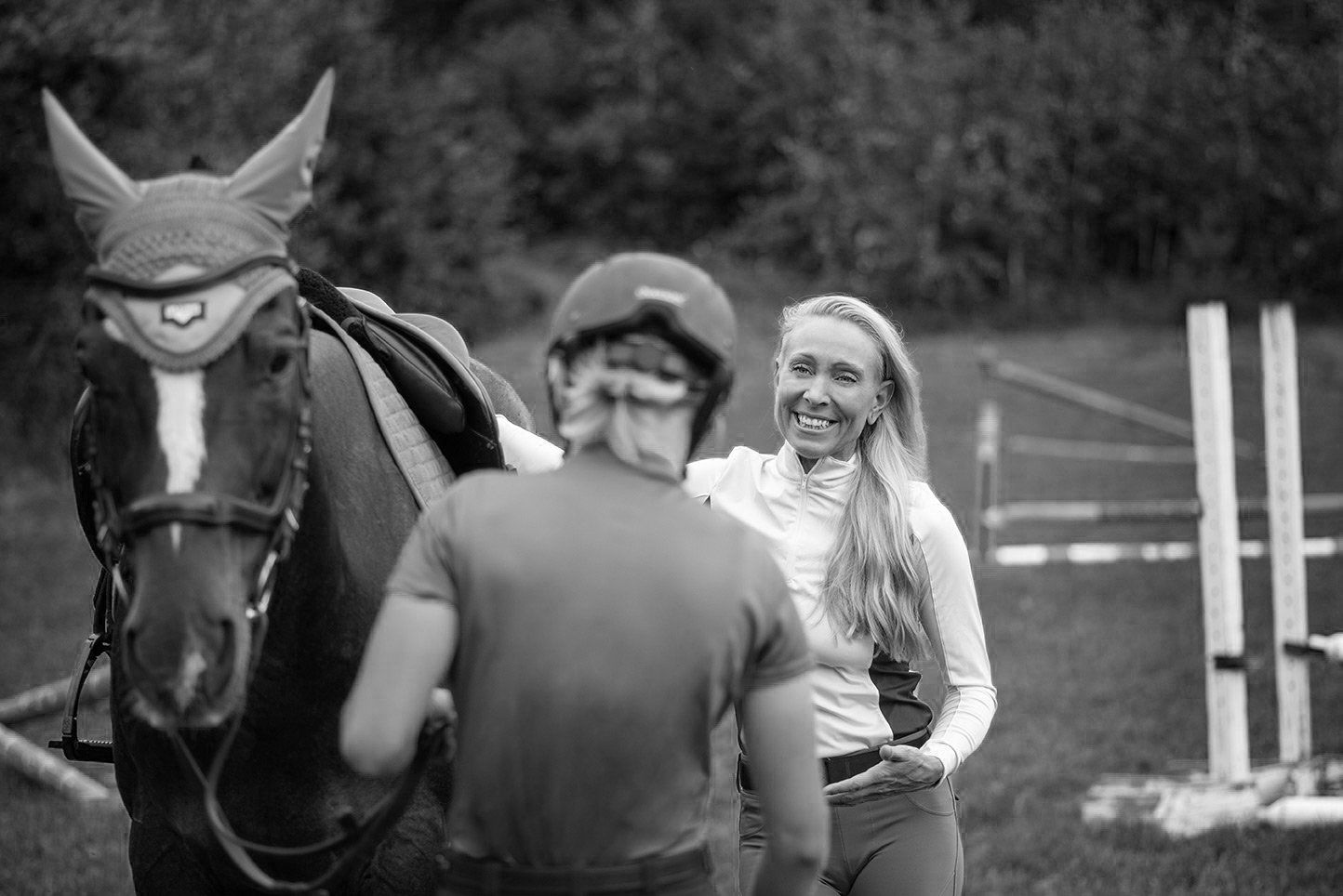 Woman smiles, stands near horse wearing a saddle and bridle, person adjusts tack. Outdoors.