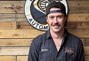 Man in black work shirt and backward cap standing before a wooden wall with a circular auto shop sign
