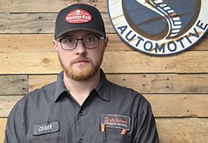Man in black shirt and cap standing before a wooden wall with automotive signs