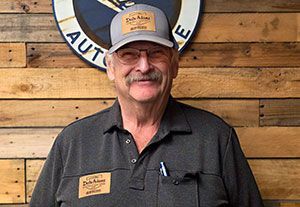 Smiling man in a gray shirt and cap standing before a wooden wall and round logo sign
