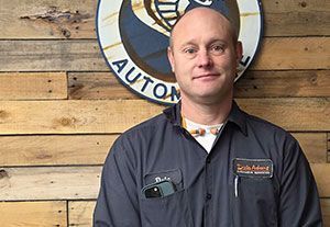 Man standing in front of a wooden wall and round auto shop sign, wearing a dark work jacket.
