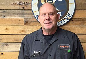 Man standing before a wood wall with a circular logo, wearing a dark work shirt and smiling
