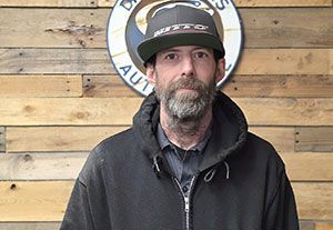 Man in a black jacket and cap standing before a wooden wall with a circular sign behind him