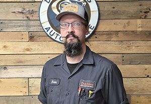 Man in glasses and cap standing before a wooden wall with a round auto shop sign.