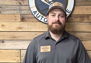 Man in gray shirt and cap standing before a wooden wall with a circular sign behind him