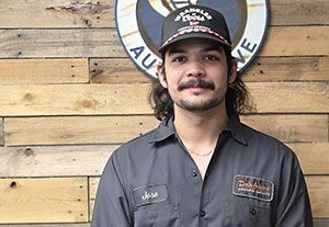 Man in a dark cap and gray shirt standing before a wooden wall and round sign, facing the camera with a slight smile