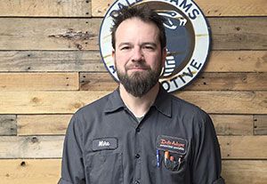 Man in a dark cap and gray shirt standing before a wooden wall and round sign, facing the camera with a slight smile