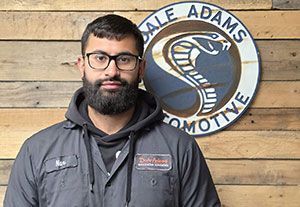 Man standing in front of a Dale Adams Automotive sign on a wooden wall, wearing a gray work jacket.