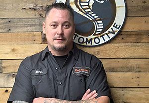Man in black work shirt stands with arms crossed in front of a wooden wall and circular logo sign