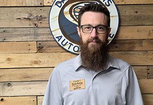 Man standing in front of a wood wall with a round auto shop sign, wearing glasses and a gray shirt.