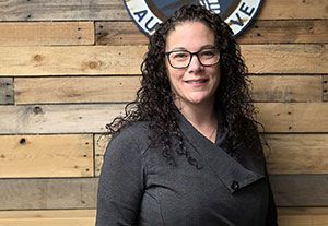 Woman with curly hair and glasses standing before a rustic wooden wall, smiling at the camera.
