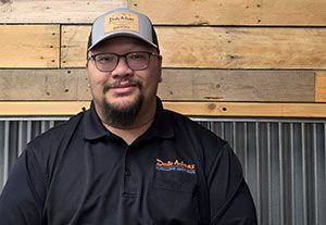 Man in glasses and cap wearing a black polo, standing against a wood plank wall and corrugated metal backdrop