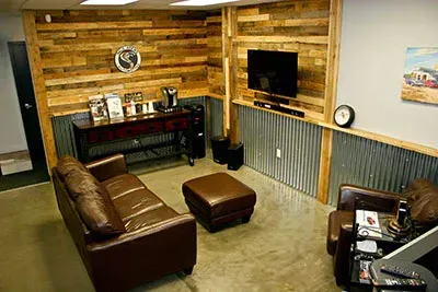 A rustic-industrial lounge with wood-paneled walls, corrugated metal wainscoting, a brown leather sofa, and a television. | Dale Adams Automotive Specialists