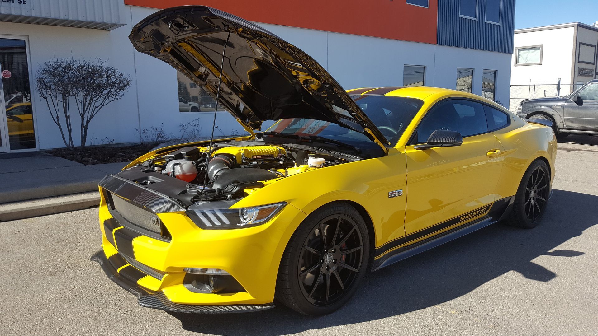 Yellow sports car with hood open, parked outdoors in front of a building.