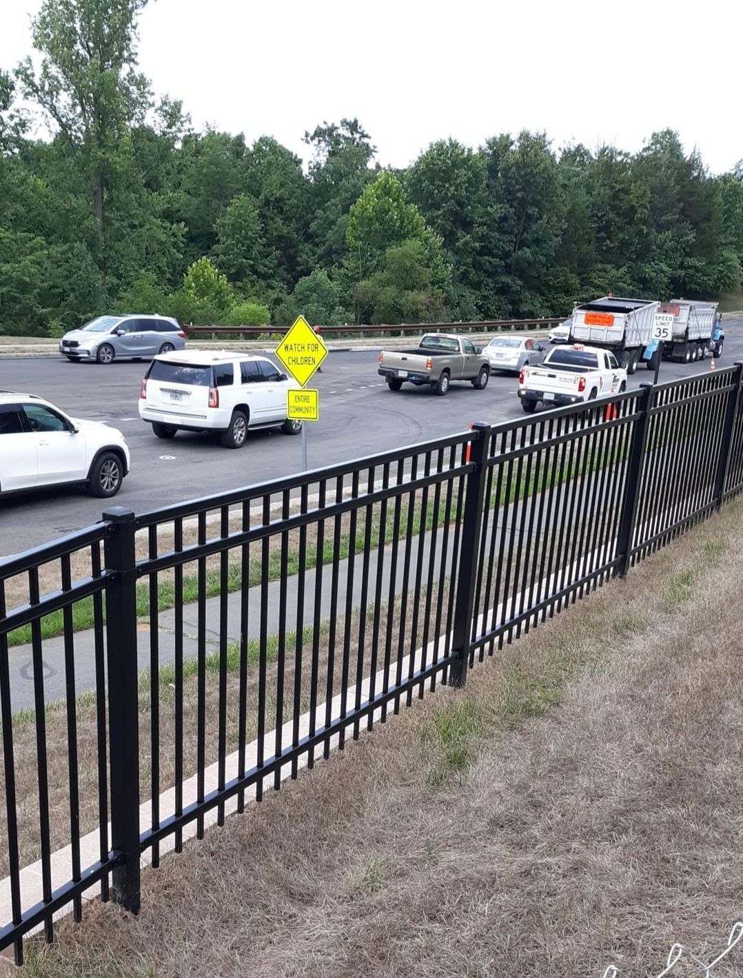 Cars traveling on a road with a black fence in the foreground and trees in the background. A yellow sign is visible.