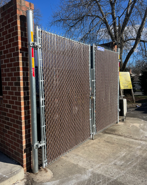 Brown chain-link fence with privacy slats, attached to a brick building. Concrete ground, sunny day.