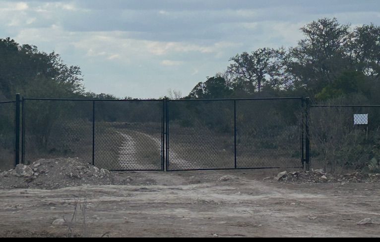 Black chain-link gate blocks dirt road leading into a wooded area under a cloudy sky.