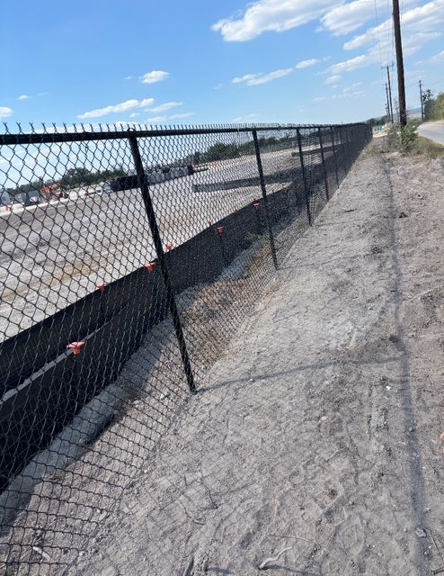 Black chain-link fence along a gravel shoulder next to a paved road, a commercial area beyond the fence.