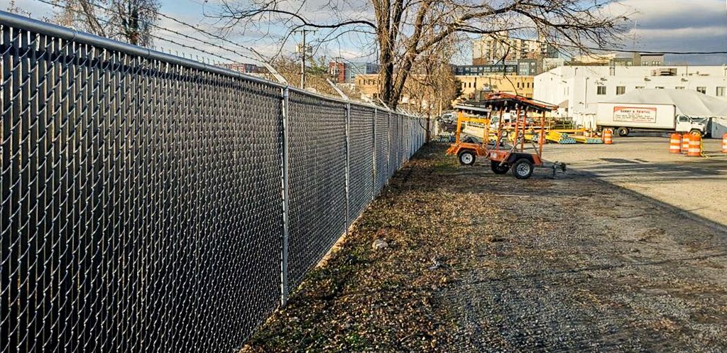 Chain-link fence topped with barbed wire runs along a grassy area, with buildings and construction vehicles visible in the background.
