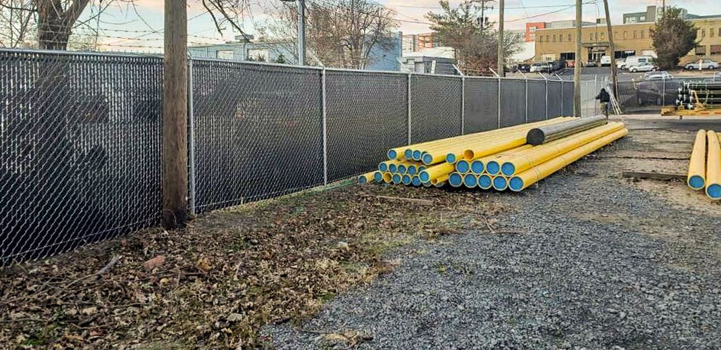 A chain-link fence borders a gravel lot with stacked yellow pipes. A distant building is visible.