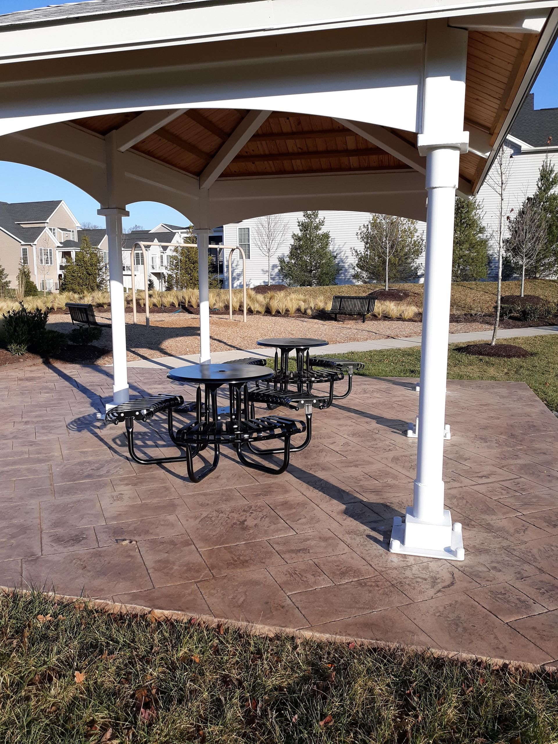 Picnic tables under a white gazebo in a park, with nearby benches and buildings in the background.