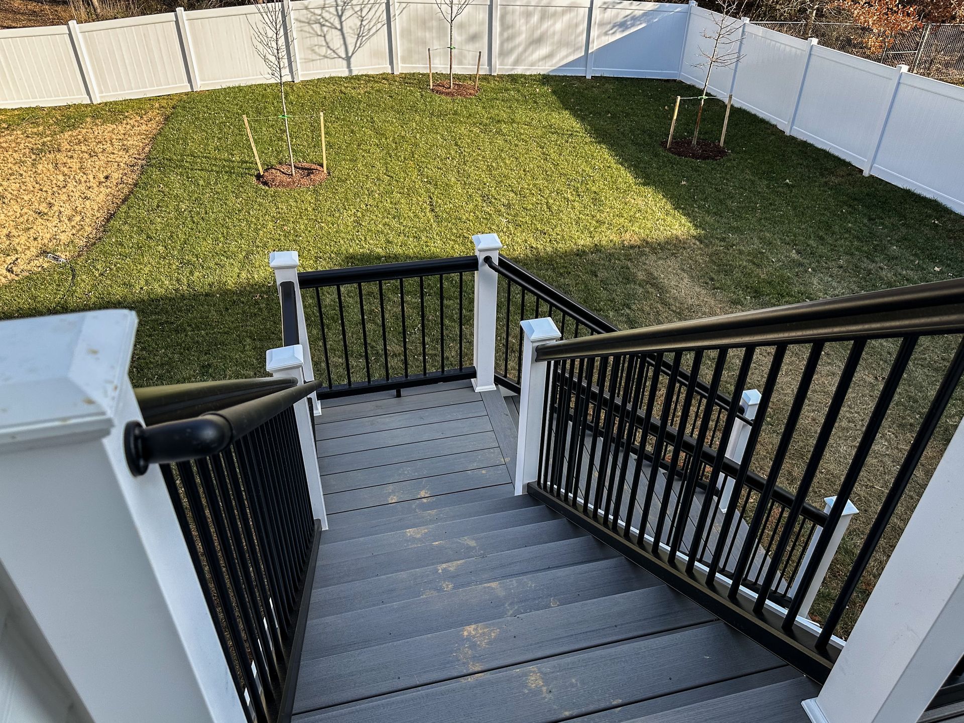 Staircase leading down to a backyard with green grass, white fence, and black and white railings.