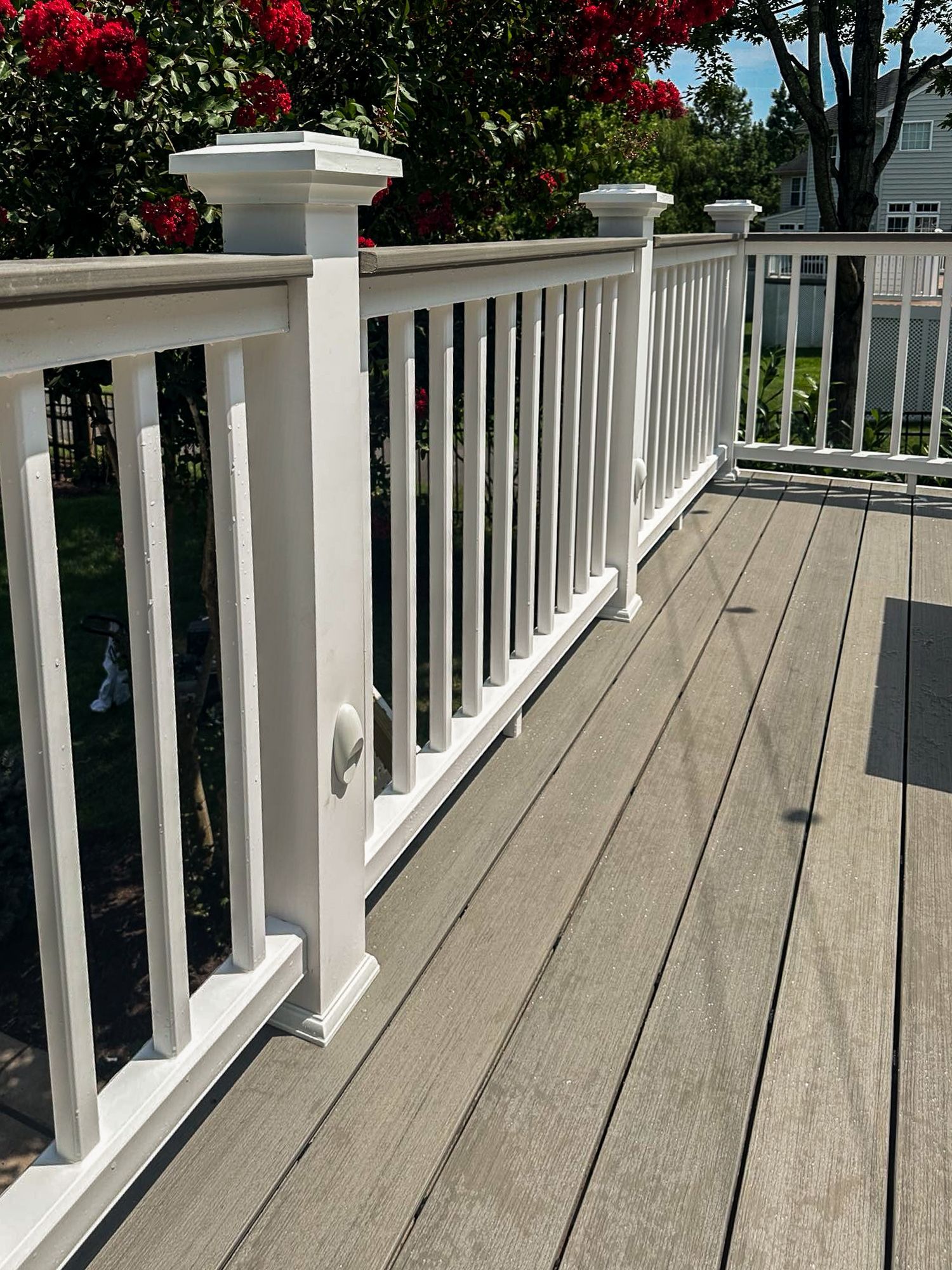 White deck railing with vertical balusters on a composite deck; lush greenery in the background.