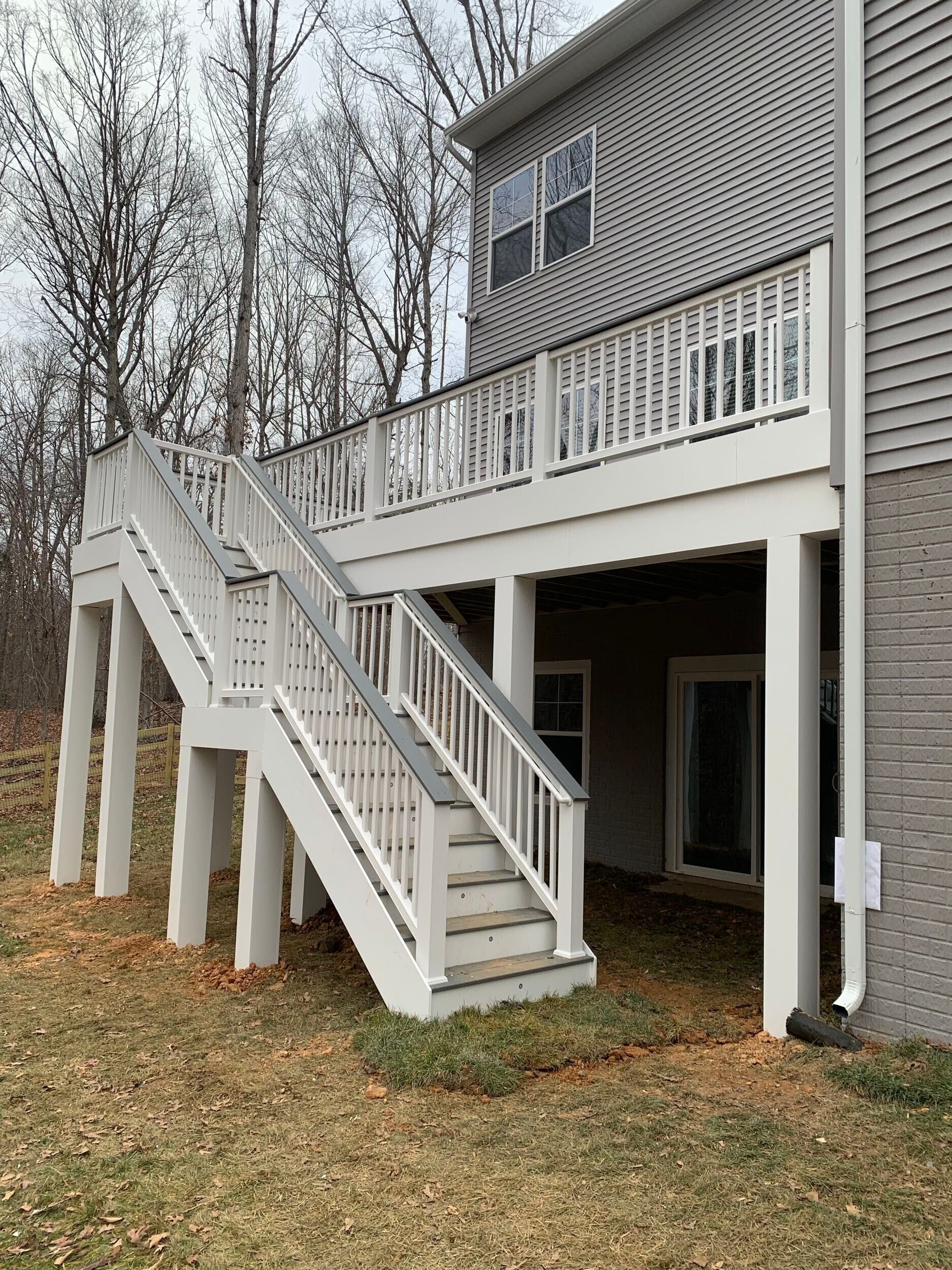White deck and stairs attached to a gray-sided house. Outdoor setting with trees and dry leaves.