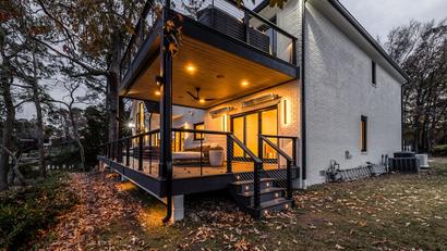 Exterior view of a modern two-story house with a lit deck and stairs, at dusk.