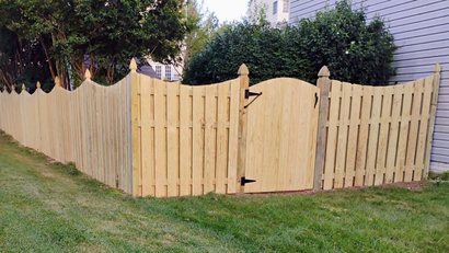 Wooden fence with gate in front yard. Light brown with black hardware. Green grass.
