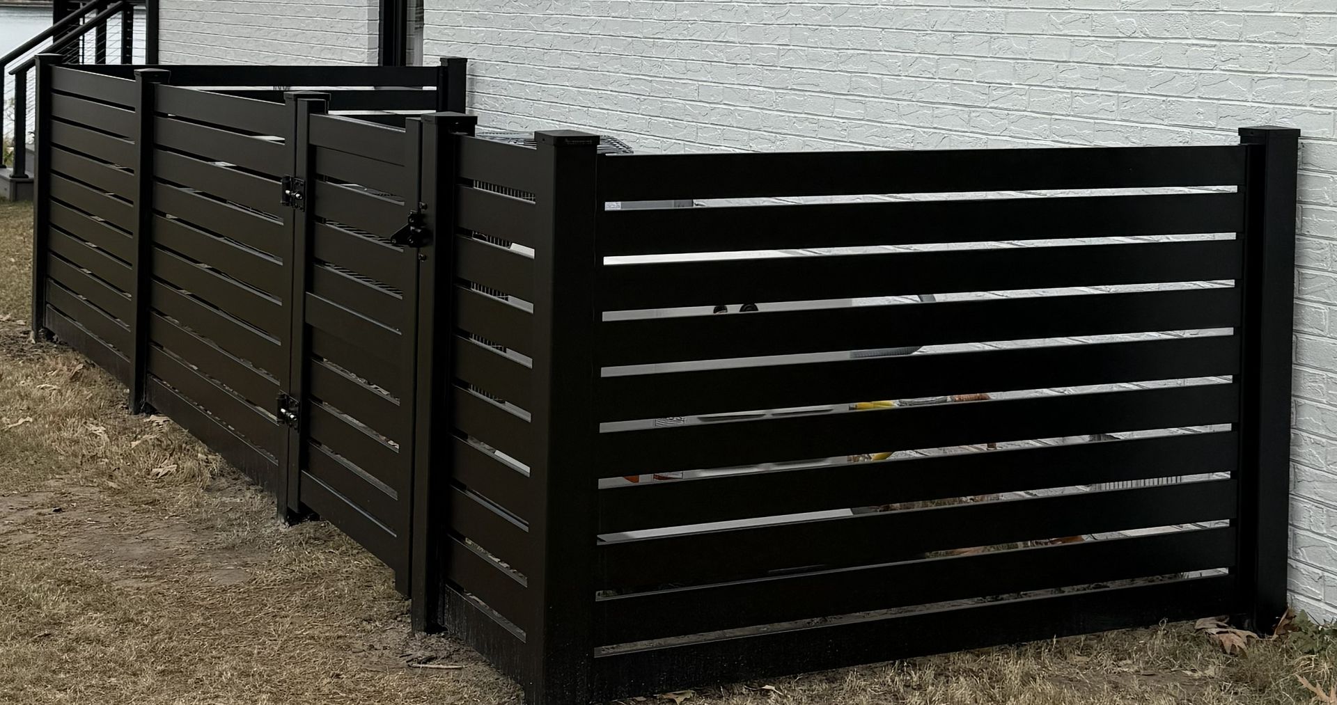 Black horizontal slat fence in a yard against a white textured wall.