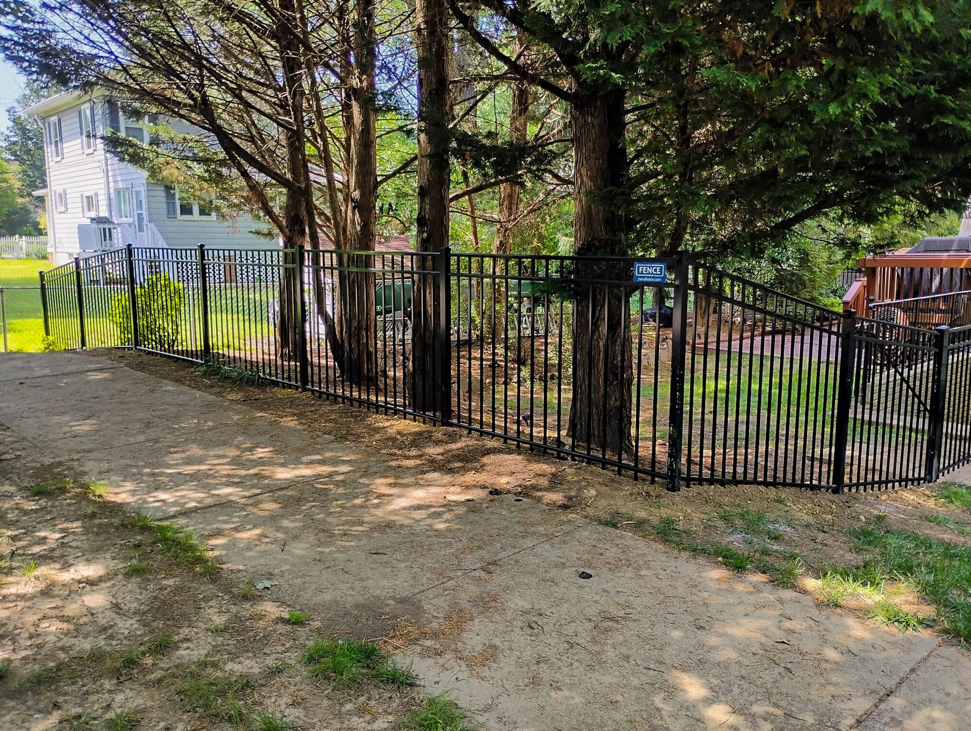 Black metal fence in front of trees, bordering a gravel path and grassy area, with a two-story house in the background.