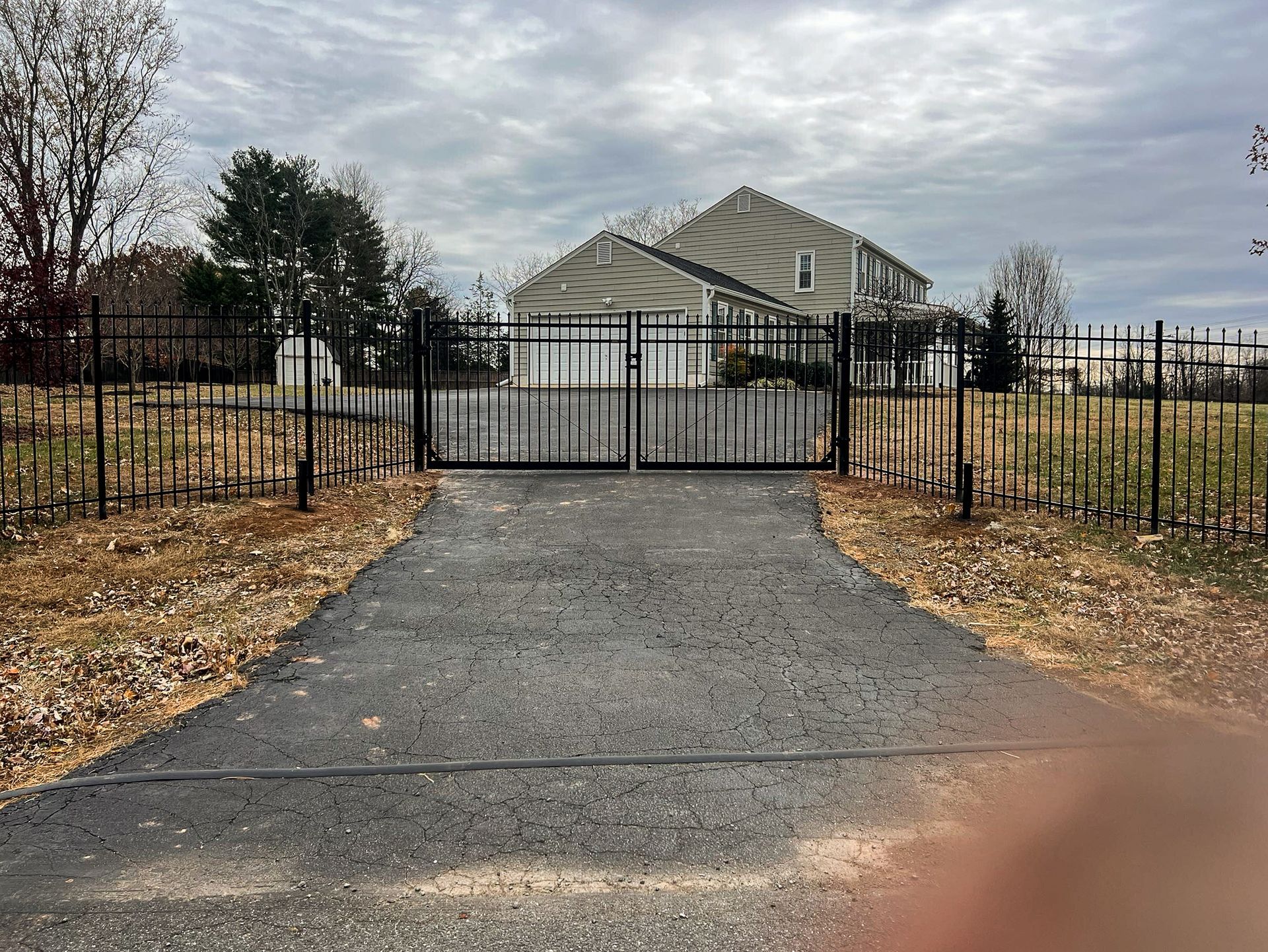 Black metal gate and fence enclose a driveway leading to a two-story house under a cloudy sky.