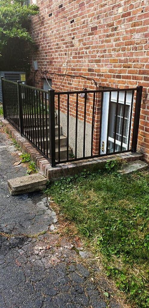 Brick building with a door at the base of stairs, enclosed by a black metal fence. Grass and asphalt surrounding.