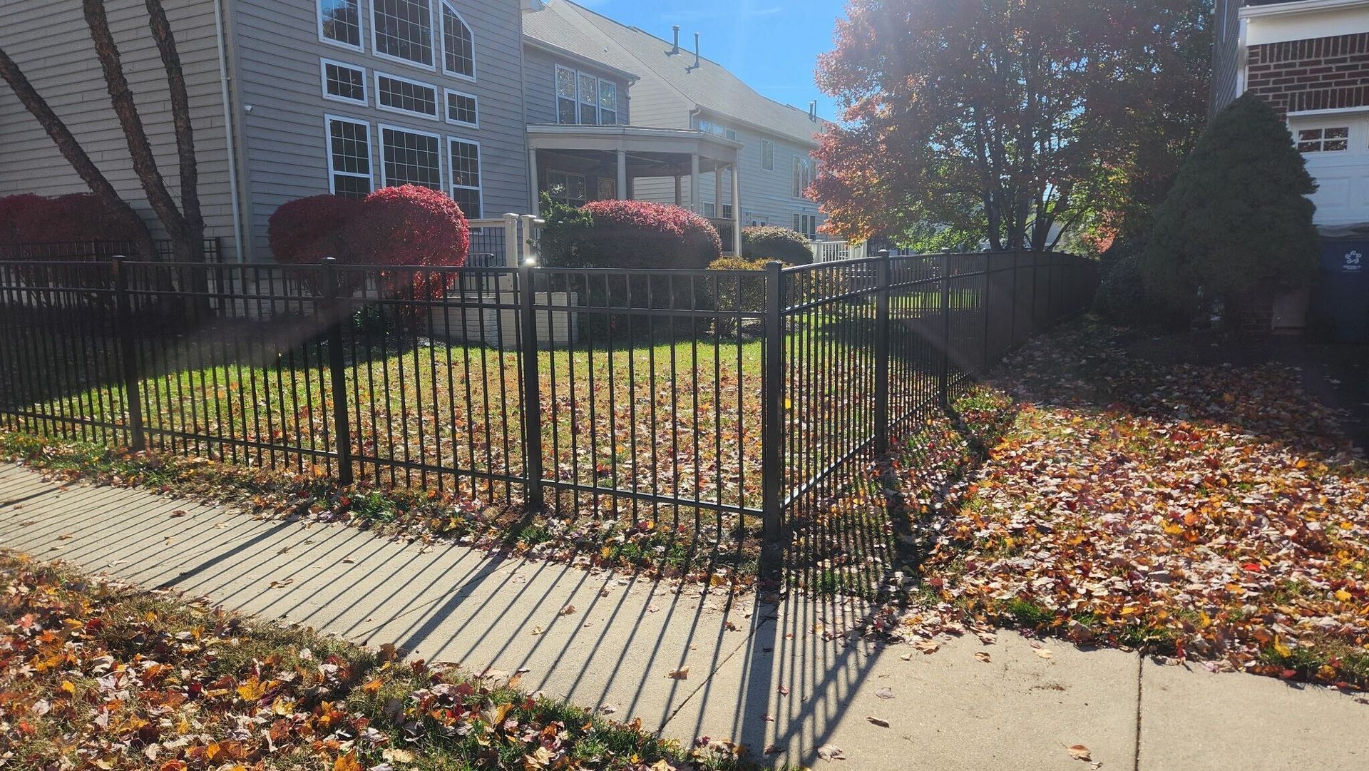 Black fence encloses a small grassy yard in front of a multi-story building. Fall leaves cover the ground.