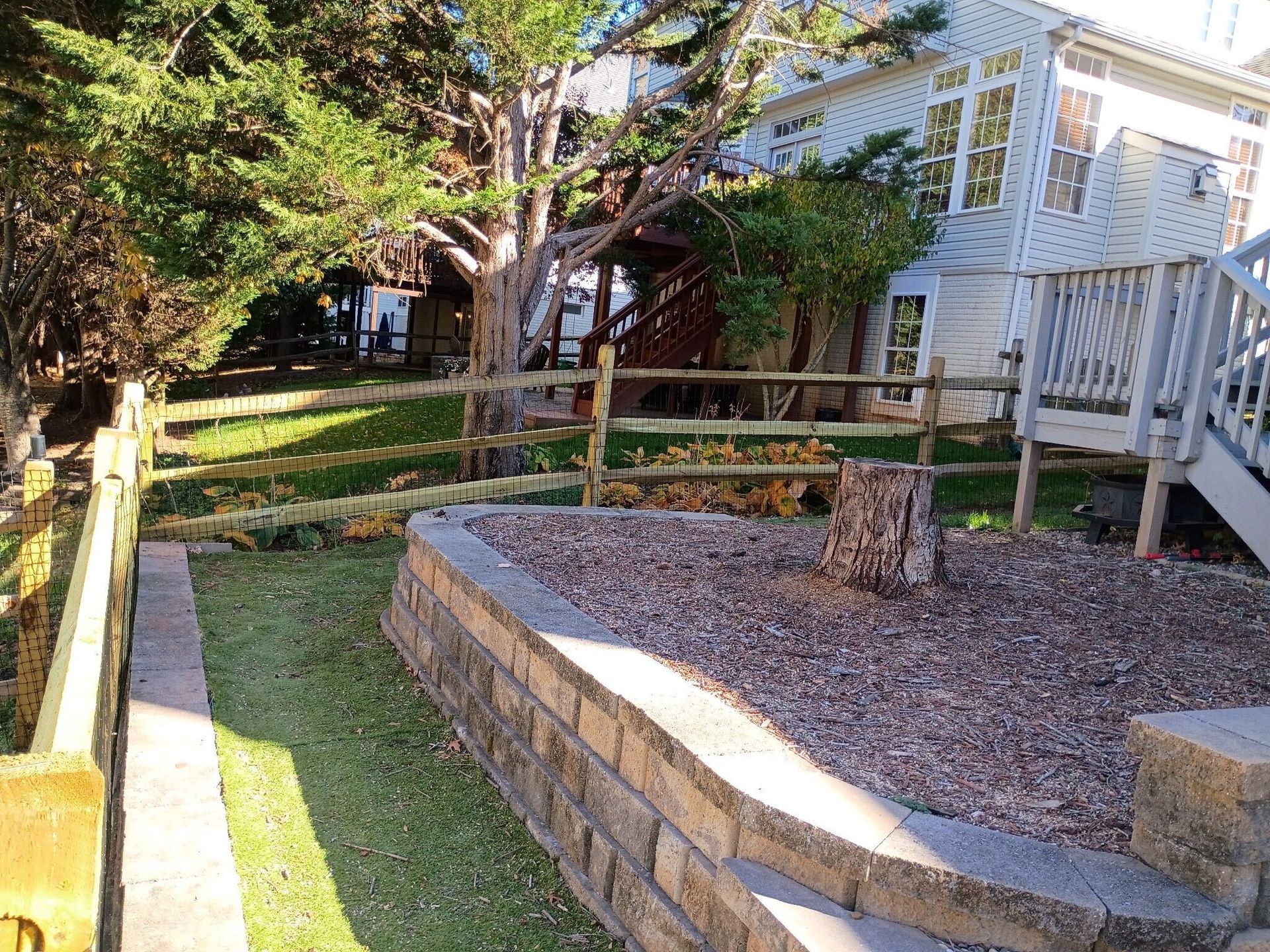 A backyard with a retaining wall, lawn, trees, and split-rail fence alongside a house.