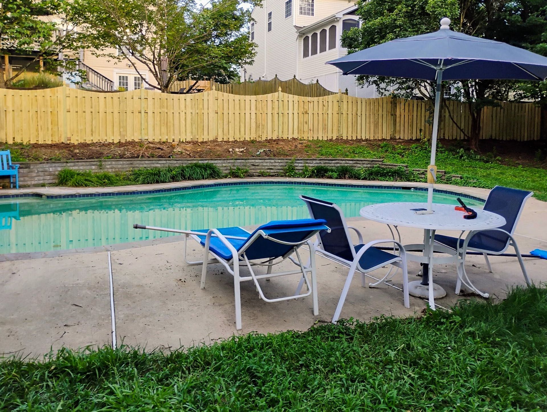 Swimming pool in backyard with patio furniture, blue umbrella, and wooden fence.