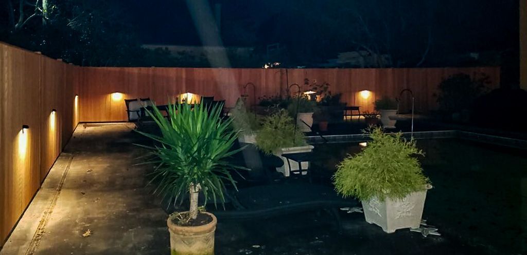 Nighttime backyard with potted plants, lit by wall-mounted lights along a wooden fence.