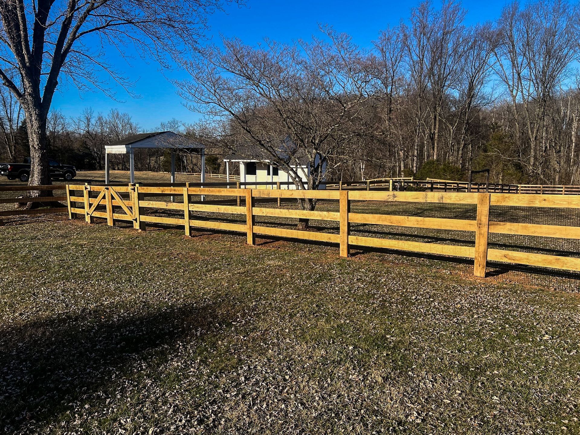 Wooden split-rail fence with gate in front of a field and small building on a sunny day.