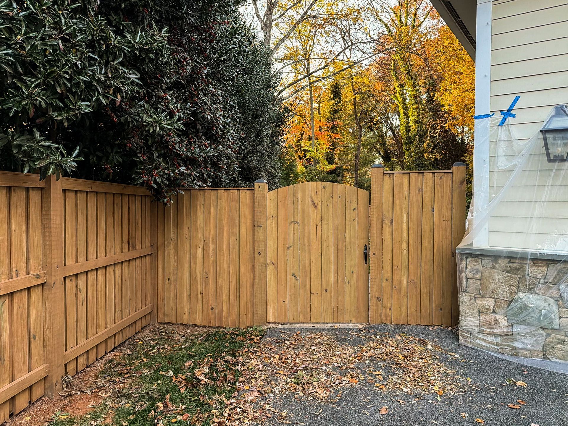 Wooden fence with a gate, autumn leaves on the ground, trees in the background, next to a house.
