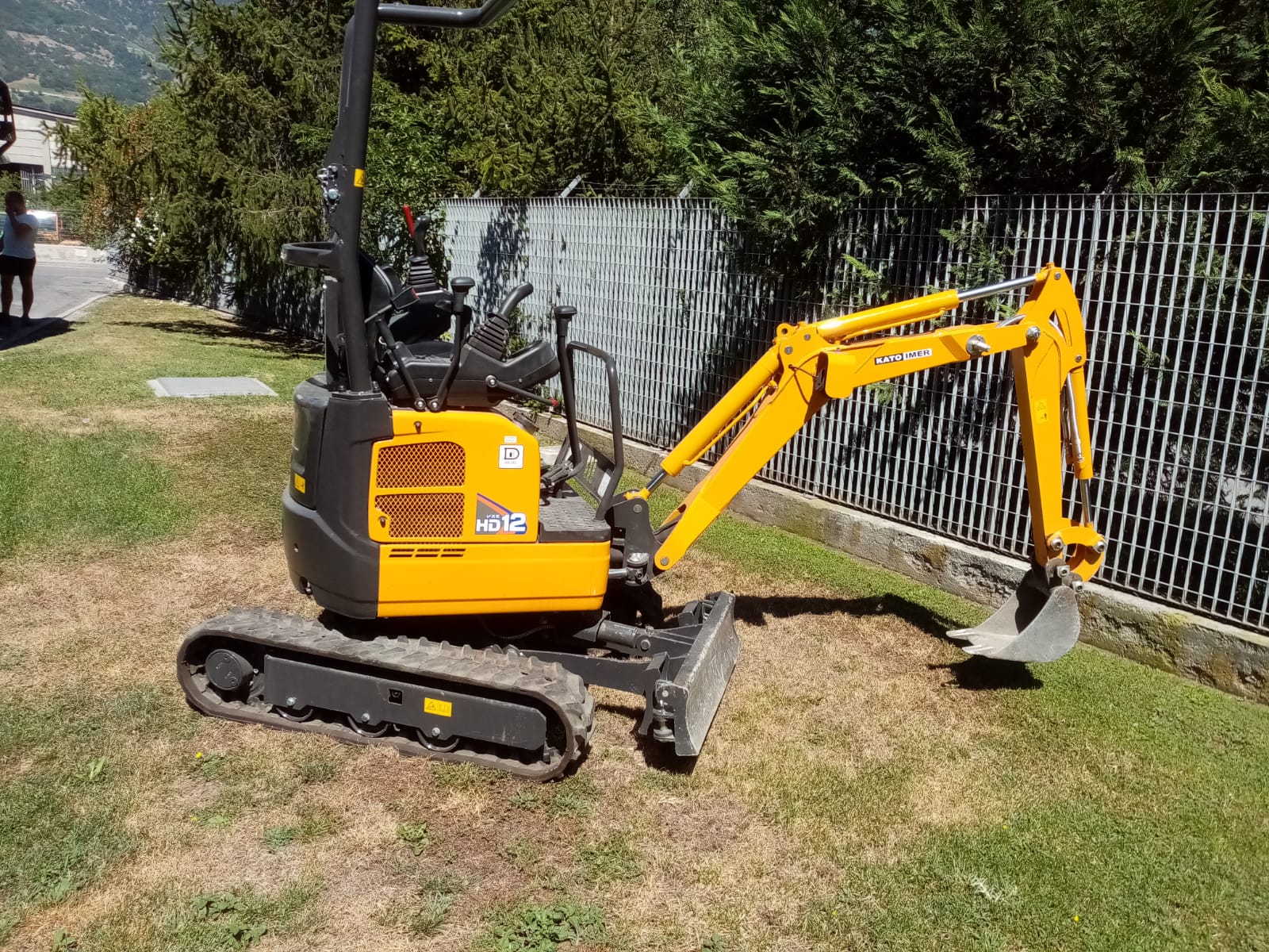 Miniescavatore giallo sull'erba accanto a una recinzione, con il braccio esteso e la benna appoggiata a terra.