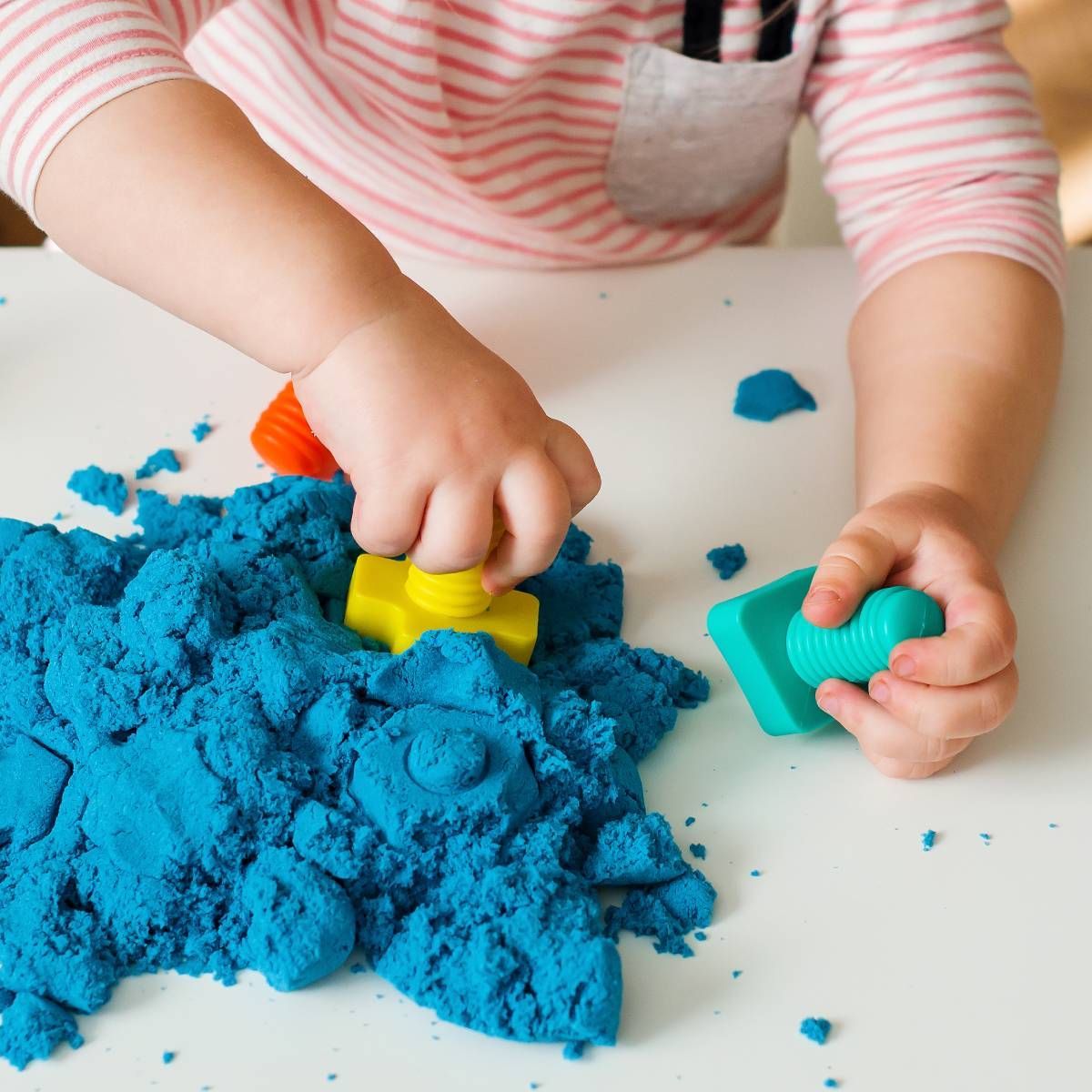 Child playing with blue kinetic sand, using colorful toy molds. White table setting.