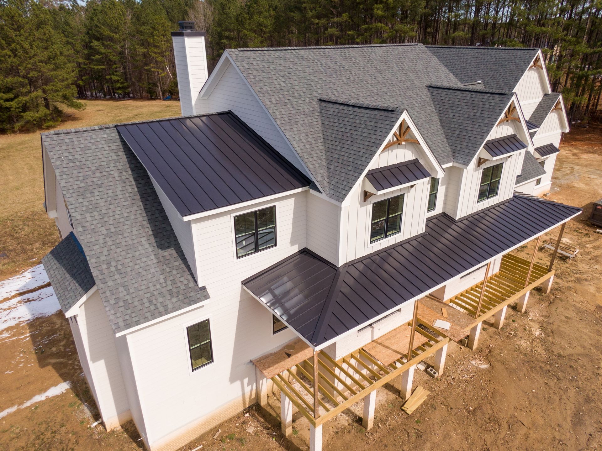 Aerial view of a new white house with gray roof and dark metal porch roof in a wooded lot