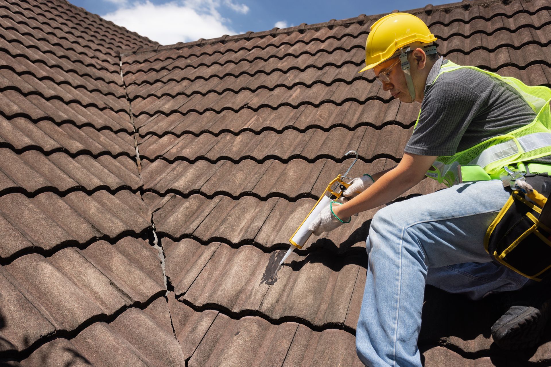 Worker in a yellow hard hat repairing a cracked tiled roof with a power tool.