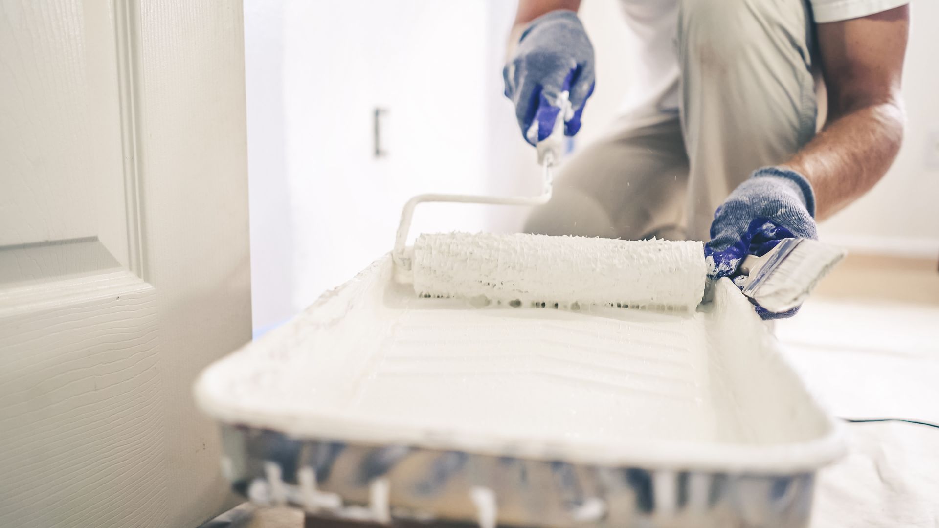 Person painting a wall with a roller tray, wearing blue gloves in a bright room